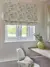 Corner of room in pale dusky pink with  a window dressed in Roman blind with flower pattern embroidered in silvers and golds, wooden desk shelf and work chair, gilt rectangular mirror on right, round yellow vase with flower pattern.