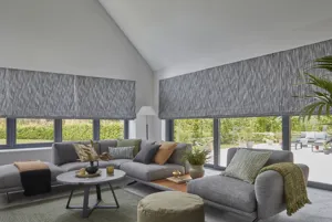 Living room with gable end and sloped ceiling windows run full length and width of both sides with grey corner sofas and chairs and windows dressed in grey marbled pattern Roman blinds.