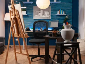 An artists studio with easeal and canvas, desk and chair and stool with a ceramic vase still life setting.