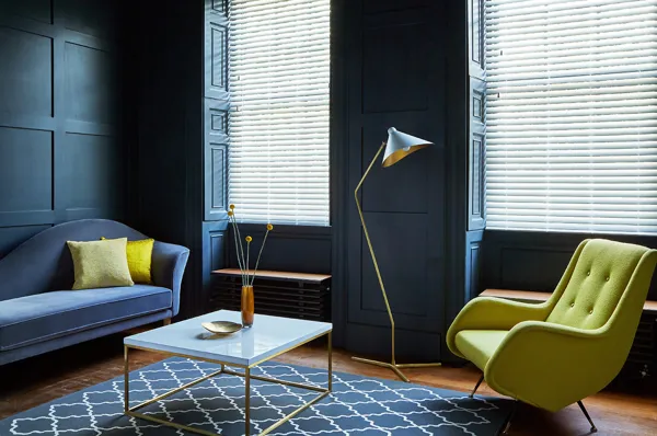 Living room with dark blue pannelled walls, two windows dressed in white wood venetian blinds, lowered with slats tilted, dark blue chaise longue with chartreuse cushions opposite a chartreuse chair wtih a blue and white diamond rug in between.