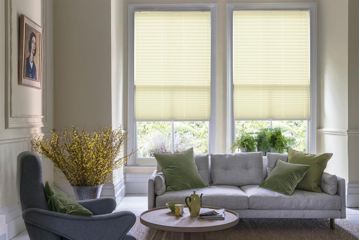 Decorated in a plain style Parchment Banana Crush yellow Pleated blinds are fitted to windows in a kitchen with marble counter tops and cream coloured fixtures.