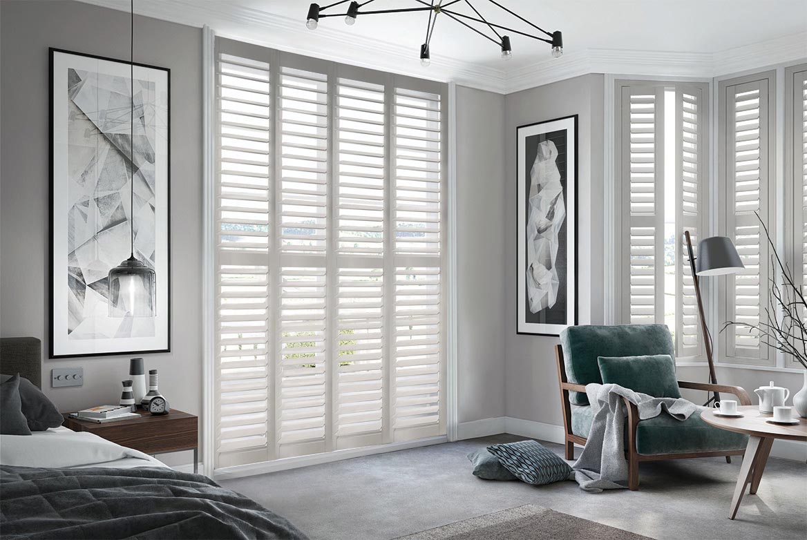 Castle Stone Shutters arranged in a tier formation are fitted to tall windows in a bedroom decorated with neutral colours of grey and white that has a bed, side table, armchair and coffee table
