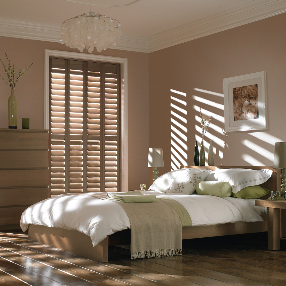Bedroom with pale mink walls, mid wood floor and furniture, white beeding  and two tall windows dressed in mid brown wooden shutters, closed with louvres open and sun rays casting on bed, wall and floors.