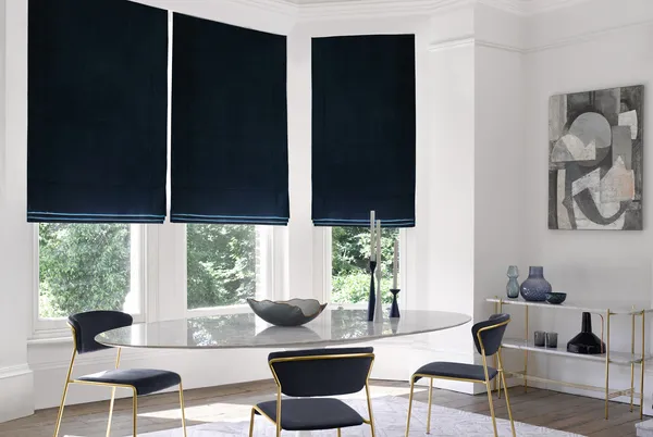 Modern dining room with bay window dressed in three navy blue velvet Roman blinds, oval glass table, metallic gold chairs with blue upholstery, metallic gold side table with blue glassware.