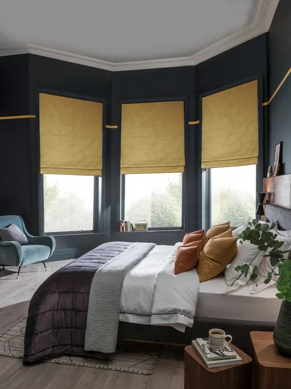 Large bedroom with bay window with three ochre faux velvet Roman blinds, wooden floor, navy walls, bed with white bedding and grey quilted throw, modern pendant light.