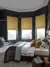Large bedroom with bay window with three ochre faux velvet Roman blinds, wooden floor, navy walls, bed with white bedding and grey quilted throw, modern pendant light.