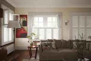 Lounge with wood floor, chocolate brown leather sofa, abstract art on walls and three sets of windows on wall to rear and left, all dressed in white tier on tier shutters, with two sets top halves open.