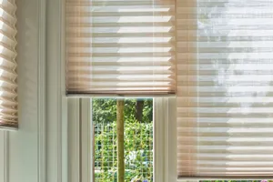 Close up of corner of living room with bay windows, dressed in pale beige pleated blinds at varying heights, brown cord sofa in window recess.