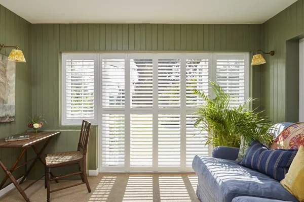 White tier on tier shutters fitted to a door window in a living room with green walls, wooden desk and blue sofas