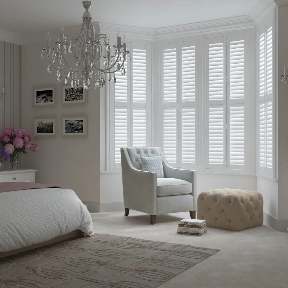 White classic-style bedroom with bay window dressed in white shutters, louvres bed on left with white bedding and dusky pink throw, white upholstered chair in bay window with pale ochre upholstered footstool, and chandelier.