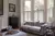 Cream lounge with bay window dressed in white birch cafe style shutters, closed with louvres open, silver grey sofa and large footstool as table, wooden side table and dark wood apothecary cabinet.