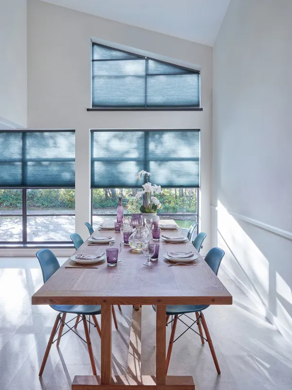 Dining area with contemporary wooden table and chairs set in front of two large windows beneath a mezzanine shaped window dressed in blue duette blinds.