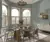 Dining room with three windows on a curved wall dressed in white cafe style shutters, closed with louvres open, pale blue walls, modern glass dining table with wood and metal chairs, white low sideboard.