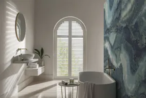 White bathroom with large arch window dressed in white shutters, closed with louvres open, abstract blue feature wall on right beside bath.