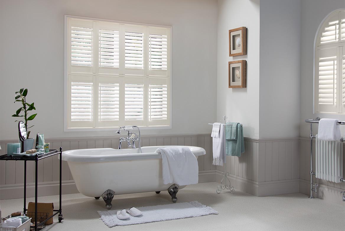 Waterberry Vanilla is a Ivory coloured Vinyl Shutter that has been fitted to a square and arch shaped window in a bathroom that has white walls, grey skirting and a bathtub in the centre of the room