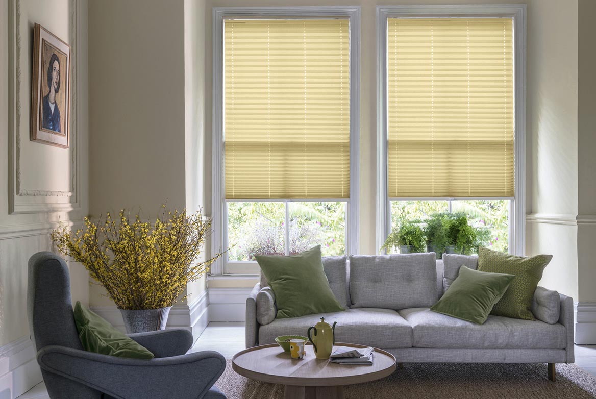 ivory Mara Shortbread Pleated blind are fitted to a series of windows in a kitchen decorated with marble counter tops, white walls and drawers. The Pleated blinds are decorated in a plain style.