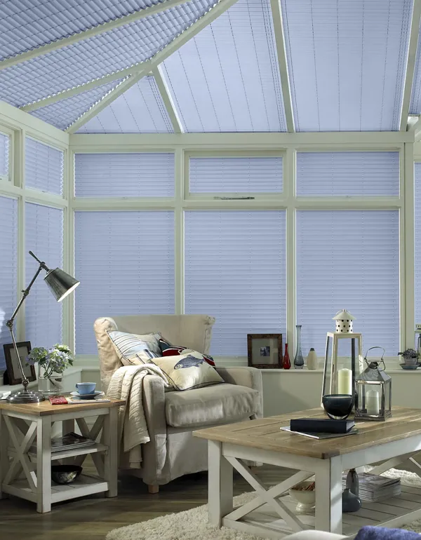 Corner of conservatory in white with pale blue side and roof blinds fully lowered, upholstered chair, white and wood coffee table and side table, hurricane lamps.