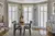 Dining room with wood floor,  three floor to ceiling windows in an extended bay dressed in white shutters, closed with louvres open, rug in grey tones, wooden table and chairs with grey cushioned seats, amber vase and greenery on table.