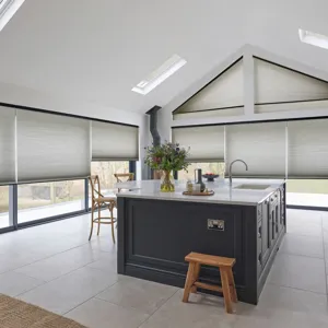 White gable end kitchen diner with white tiled floor, island area with blue units and white worktop, three skylights,  three landscape windows to left and gable end windows and doors all dressed in mid grey duette blinds, skylight blinds three quarters lowered, side blinds at varying heights, gable shape fully lowered and door blinds lowered at several heights.