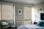 Pale grey bedroom with two windows dressed in pale stone-grey coloured wood venetian blinds, lowered, louvres tilted. bed dressed in pale blue bedding, wooden chair in corner with blue cushions.