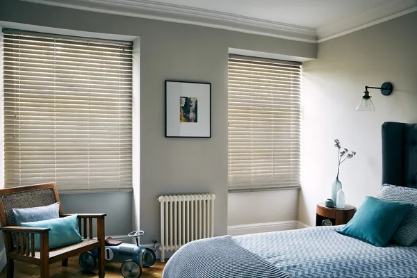 Pale grey bedroom with two windows dressed in pale stone-grey coloured wood venetian blinds, lowered, louvres tilted. bed dressed in pale blue bedding, wooden chair in corner with blue cushions.