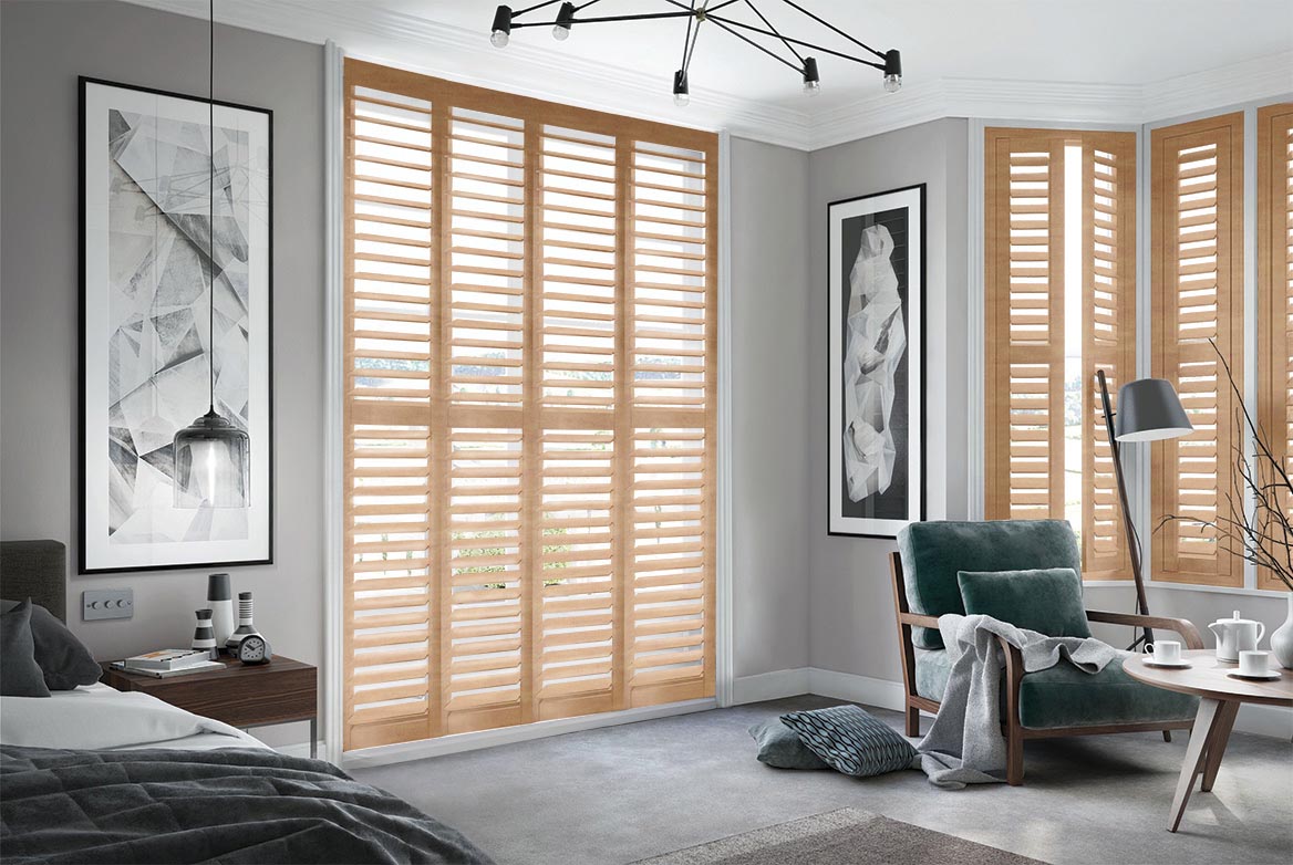 Burley Golden Fall Shutters have been fitted to tall windows in a bedroom. The Shutters are Natural, Brown coloured while the bedroom is decorated with grey tones.