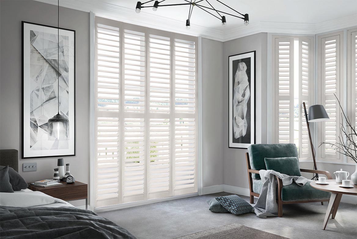 Feather Touch Shutters arranged in a tier formation are fitted to tall windows in a bedroom decorated with neutral colours of grey and white that has a bed, side table, armchair and coffee table