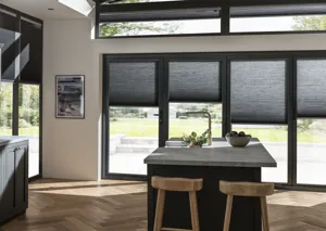 Gable end kitchen with dark grey units on left, island with marble grey workstop and sink in centre and gable end windows and doors dressed in dark grey duette blinds at varying heights.