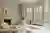 Neutral living room with bay window dressed in cream full height shutters, closed with louvres open, modern cream chairs, round low table on a abstract patterned rug in shades of cream and brown, wooden low coffee table sits in the bay alcove.