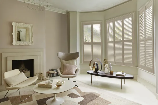 Neutral living room with bay window dressed in cream full height shutters, closed with louvres open, modern cream chairs, round low table on a abstract patterned rug in shades of cream and brown, wooden low coffee table sits in the bay alcove.