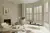 Neutral living room with bay window dressed in cream full height shutters, closed with louvres open, modern cream chairs, round low table on a abstract patterned rug in shades of cream and brown, wooden low coffee table sits in the bay alcove.