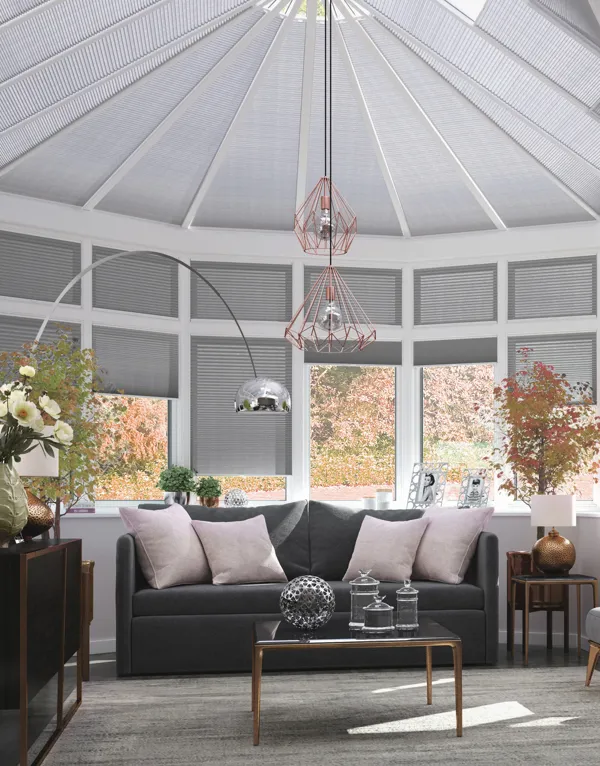Extended conservatory with side and roof windows dressed in pale grey duette blinds.grey flooring, grey rug, dark grey upholstered sofa and chairs, low table with glassware, pale pink cushions, low-hanging pendant light with wirework shades.