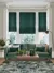 Pale grey lounge with pale wood floor and large faded geometric patterned forest green rug with table and green accessores, green sofa with cushions in dark green and cream in front of bay window dressed in two narrow and one wide Roman blinds in deep green with feint jacquard weave