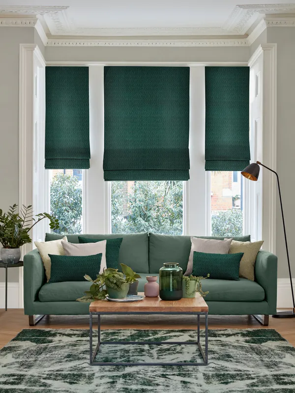 Pale grey lounge with pale wood floor and large faded geometric patterned forest green rug with table and green accessores, green sofa with cushions in dark green and cream in front of bay window dressed in two narrow and one wide Roman blinds in deep green with feint jacquard weave
