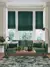 Pale grey lounge with pale wood floor and large faded geometric patterned forest green rug with table and green accessores, green sofa with cushions in dark green and cream in front of bay window dressed in two narrow and one wide Roman blinds in deep green with feint jacquard weave