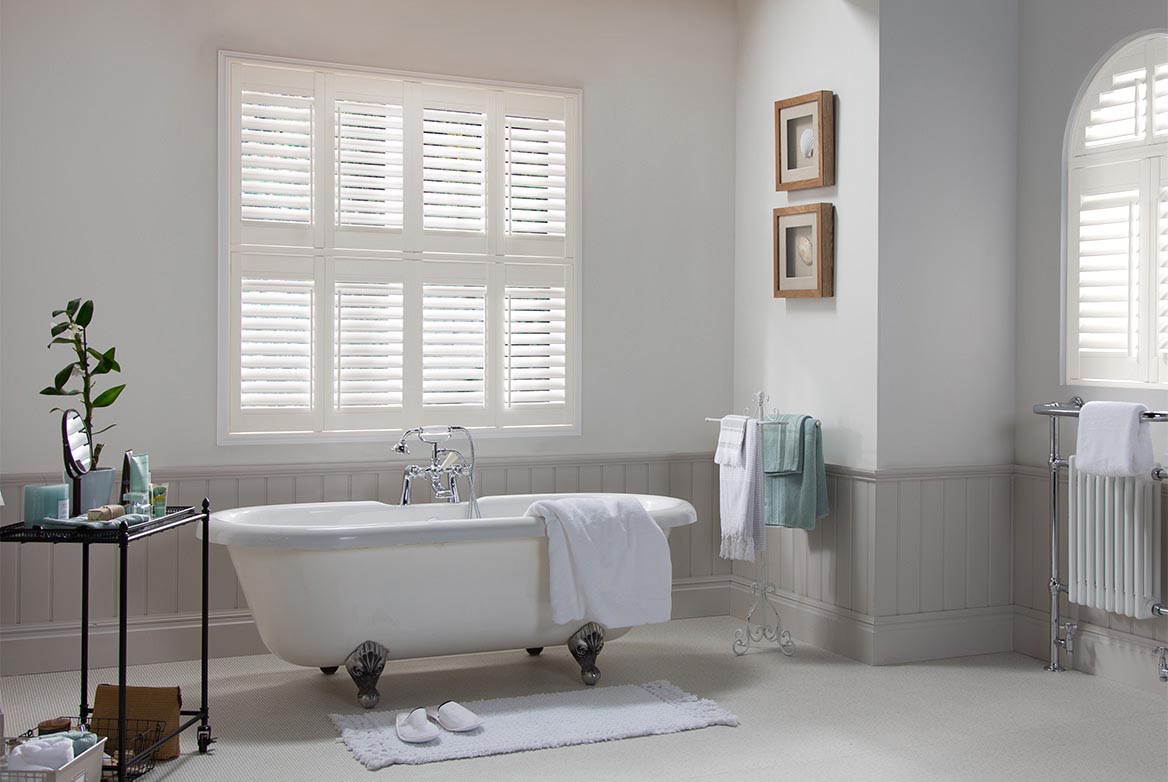 White decorated bathroom with a bathtub in the centre has Waterberry Ivory Vinyl Shutters fitted to a square and arch shaped window in the room. The Vinyl Shutters are in a Ivory colourway