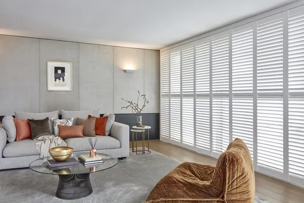  Grey ounge with full wall of white tracked shutters floor to ceiling louvres open, grey sofa terracotta cushions background, upholstered gold velvet chair foreground, a glass circular coffe table in between.