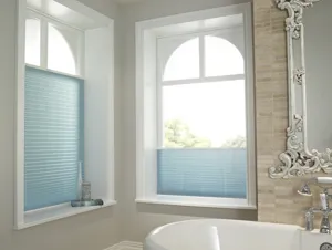 Corner of bathroom with two windows either side dressed in pale blue pleated blinds raised up from the bottom, bath beneath a gilt mirror.