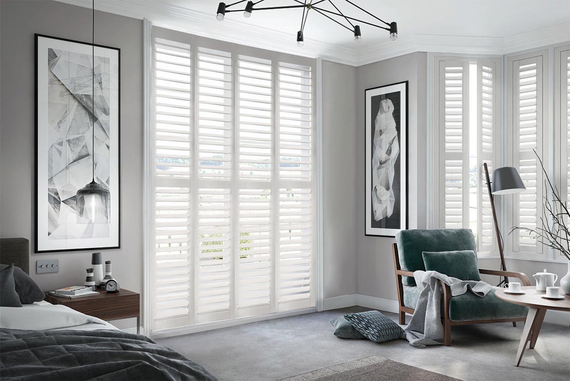 In a bedroom that has a bed, sidetable, armchair and coffee table are Ginberry Blossom Shutters. The Ivory coloured Shutters are fitted to a series of tall windows in the bedroom which is decorated in neutral grey tones.