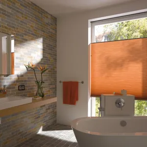 Bathroom with picture window in white wall, dressed in a cadmium orange blind in the middle of the window, side wall and floor tiled in metro stile in shades of brown, grey and copper, white bathtub in centre, pale wood shelf with white sink, cadmium orange towel on rail by window.