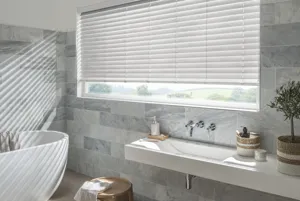 White bathroom with wide window with shallow drop, dressed in stone-coloured faux wood venetian blind, lowered three quarters, tilted with rays of light coming through, tiling to walls from floor up to three quarters high in shades of grey, white bath on raised platform, white shelf with integrated sink under window, brown tiled floor.