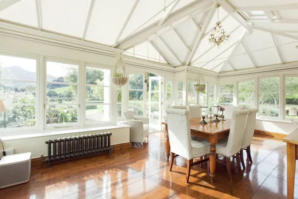 Large conservatory with white duette blinds to roof, wooden floor, dining table and chairs in centre, and views over the garden and hills..