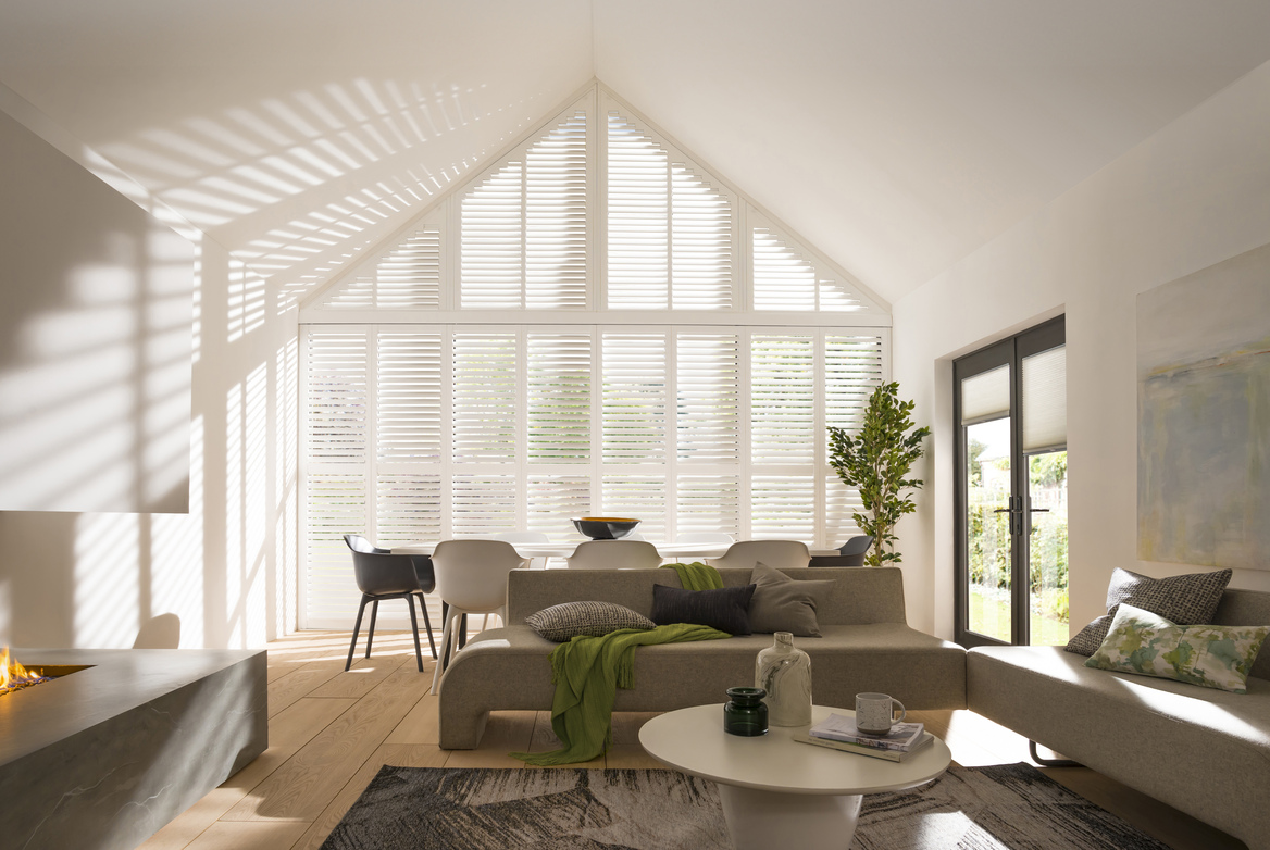 Large open-plan living-diner with gable end windows and doors dressed in white shutters, feature fireplace, lit, on left