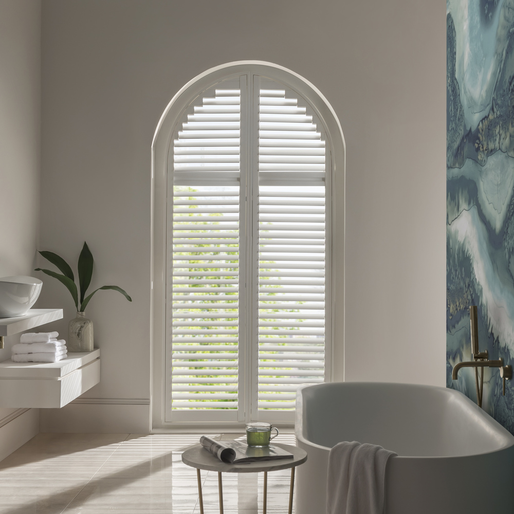 White bathroom with large arch window dressed in white shutters, closed with louvres open, abstract blue feature wall on right beside bath.