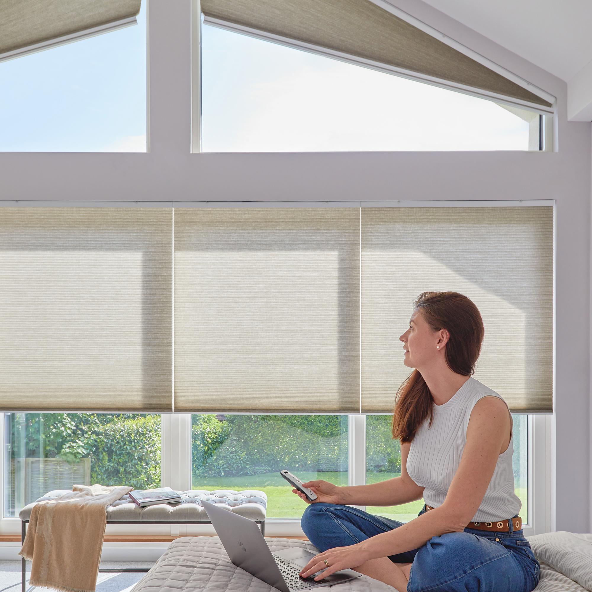 Pale grey bedroom with gable end windows dressed in pale beige duette blinds, top shaped blinds raised almost totally, lower blinds halfway, a woman with a laptop sits on a bed with a remote control for the blinds in her hand.