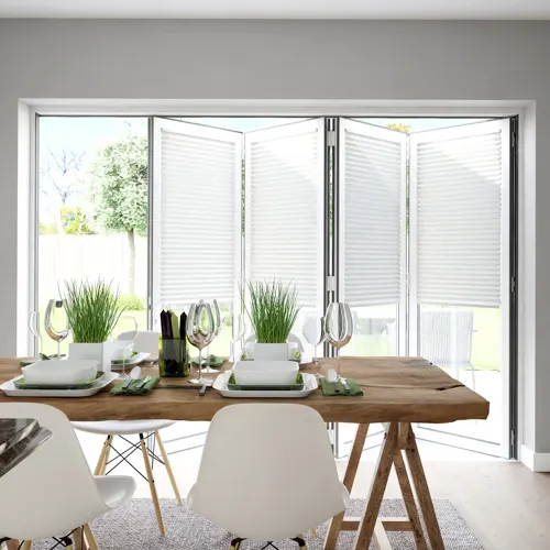 Dining room in pale grey with pale wood floor, wooden trestle-style table with white contemporary chairs, table set with white crockery and bifold doors dressed with white blinds within frame, doors partially open.