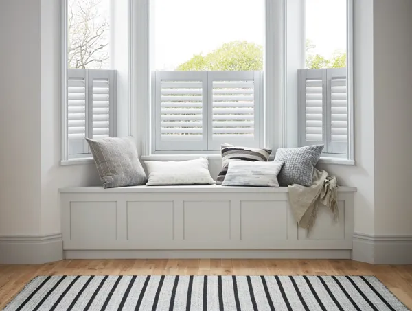 White room with wooden floor, white and blue striped rug in front of a window seat with pale blue and white cushions and cafe style shutters to window, in white.