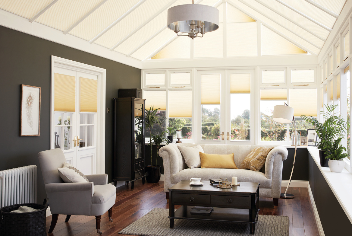 Conservatory with dark wood floor, cream sofa and chairs, dark wood coffee table with windows, doors and roof dressed in ivory duette blinds, window blinds at varying heights with roof blinds closed..