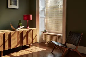 Pale wooden venetian blind in a tall narrow window, fully lowered with slats open in a green  living room.