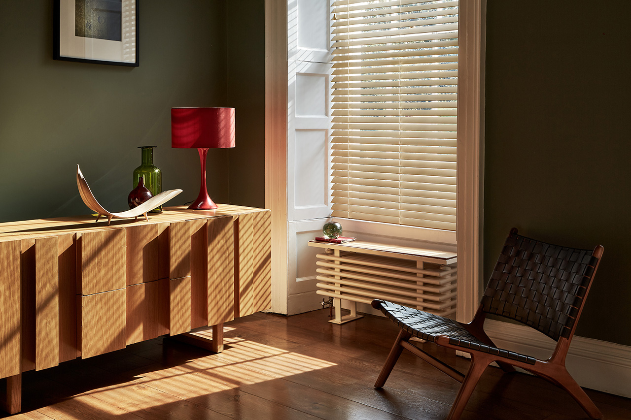 Pale wooden venetian blind in a tall narrow window, fully lowered with slats open in a green  living room.